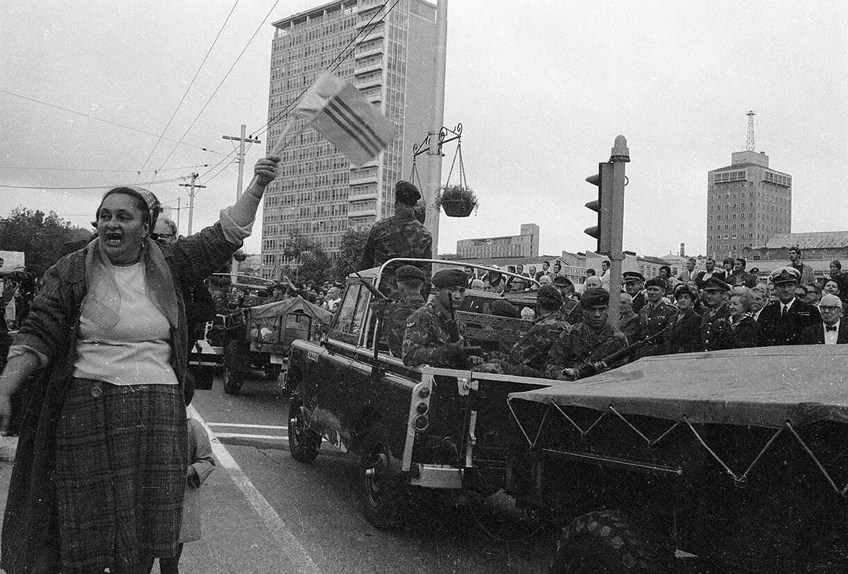 Aunty Helen, ‘Aunty’ Helen Kesha of Ngati Whatua, flourishing South Vietnamese Government paper flag, welcoming the SAS contingent in the 161 Battery parade, while hurling abuse at the protest contingent beyond, 23 May 1971